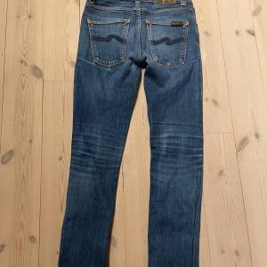Blå jeans från Nudie Jeans - Säljer ett par klassiska blå jeans från Nudie i modellen Grim Tim. Storlek 27/30. Jeansen har en liten defekt men går lätt att lag och syns knappast. 