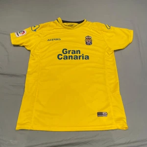 Gul fotbollströja Las Palmas Gran Canaria - Säljer en gul fotbollströja från Las Palmas med Gran Canaria-tryck på bröstet och klubbmärke. Tröjan har korta ärmar och blå detaljer. Perfekt för dig som vill visa ditt stöd för laget!
