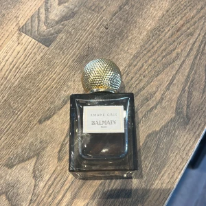 Ambre Gris parfym från Balmain - Lyxig parfymflaska från Balmain med doften Ambre Gris. Flaskan är fyrkantig i mörkgrått glas och har en rund, guldig kork med textur. Elegant design som passar perfekt på parfymhyllan.