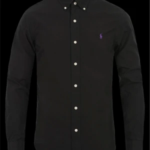 Svart skjorta från Polo Ralph Lauren - Stilren svart skjorta från Polo Ralph Lauren med smal passform och klassisk knappslå. Skjortan har vita knappar och en lila broderad logga på bröstet. Perfekt för dig som gillar en clean och tidlös look.