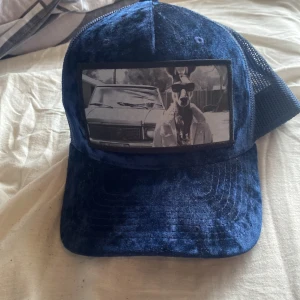 Blå sammetskeps med mesh och tryck - Snygg blå keps i sammetsmaterial med svart mesh baktill och justerbar knäppning. Framtill finns ett svartvitt tryck med motiv av en hund i cowboyhatt framför en bil. Perfekt för dig som vill sticka ut.