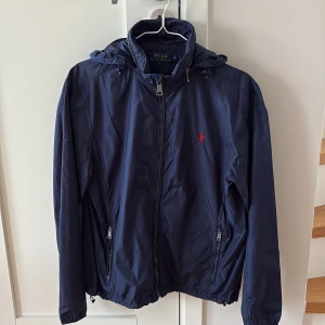 Mörkblå vindjacka från Polo Ralph Lauren - Säljer en mörkblå vindjacka från Polo Ralph Lauren med huva och dragkedja framtill. Jackan har två fickor med dragkedja och en liten röd logga på bröstet. Perfekt för blåsiga dagar och enkel att matcha med det mesta.