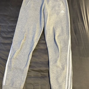 Grå mjukisbyxor från Adidas med vita ränder - Säljer ett par grå mjukisbyxor från Adidas med klassiska vita ränder längs sidorna och logga på höften. Byxorna har resår i midjan och vid bensluten, perfekt för chill eller träning. Byxorna är oanvända.