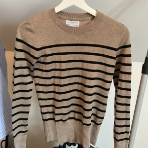 Beige och svart randig tröja från Holly & Whyte - Säljer en beige stickad tröja från Holly & Whyte med svarta ränder. Tröjan har långa ärmar, rund halsringning och dekorativa knappar vid ärmsluten. Perfekt för en stilren och klassisk look. Aldrig använd🥰