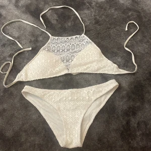 Vit spetsbikini med knyt - Säljer en söt vit bikini med spetsdetaljer och knyt i nacken. Bikinin har ett högre halterneck-liv och matchande trosor med samma spetsmönster. Bikinin har storlek S i trosorna och L i toppen
