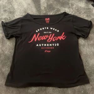 Svart t-shirt med New York-tryck - Svart t-shirt med rund hals och tryck i vitt och rött framtill: 'Sports Wear New York Authentic Division'. T-shirten har korta ärmar och en avslappnad passform. Perfekt vid träning!