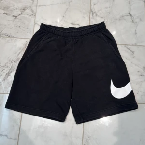 Svarta shorts från Nike med vit logga - Svarta shorts från Nike med stor vit Swoosh-logga på ena benet. Shortsen har resår i midjan och sidofickor, samt en bakficka. Perfekta för träning eller avslappnade dagar.