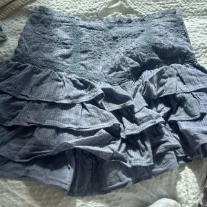 Duvblå volangkjol med broderi - Supersöt grå kjol med flera volanger och broderade detaljer upptill. Kjolen har en kort modell och ger en romantisk vibe med sina lager och struktur. Perfekt att styla med en enkel topp eller stickad tröja.