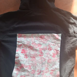 Svart hoodie med tryck - Säljer en svart hoodie med en stor ficka fram och en unik tryckdetalj på ryggen i rosa och vitt. Perfekt för en avslappnad stil. Huvan har dragsko för justering. Har sytt den själv