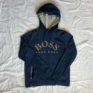 Mörkblå hoodie från Hugo Boss - Säljer en mörkblå hoodie från Hugo Boss med stor logga framtill. Tröjan har huva med dragsko och långa ärmar. Perfekt för en avslappnad och stilren look.