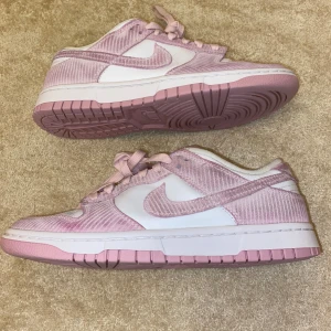 Nike Dunk sneakers i rosa och vitt - Säljer ett par Nike Dunk sneakers i rosa och vitt med snörning. Skorna har en låg modell med vit bas och rosa paneler i ett ribbat material. Sulan är rosa och vit, och Nike-loggan syns tydligt på sidan. Perfekta för dig som gillar snygga och trendiga sneakers.