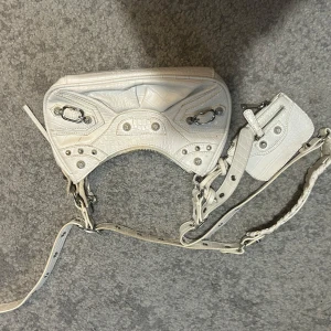 Vit axelväska  - Säljer en vit axelväska  med silverfärgade metalldetaljer och dragkedja framtill. Väskan har justerbar axelrem och en liten avtagbar spegel. Perfekt för dig som vill ha en stilren och trendig accessoar.