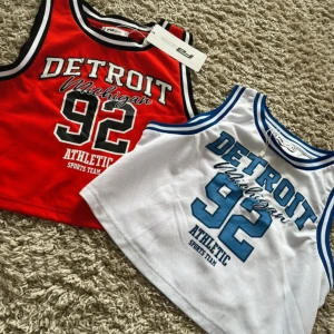 Basketlinne Detroit Michigan 92 från FB Sister - Säljer två basketlinnen från FB Sister med tryck 'Detroit Michigan 92'. Ett är rött med svarta och vita detaljer, det andra är vitt med blå detaljer. Båda har sportig look och är ärmlösa med rund hals.