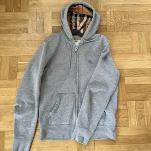 Grå hoodie med dragkedja från Burberry - Säljer en grå hoodie från Burberry med dragkedja och klassiskt rutigt foder i huvan. Tröjan har två fickor framtill och ribbade muddar vid ärmslut och nederkant. Perfekt för en avslappnad stil.