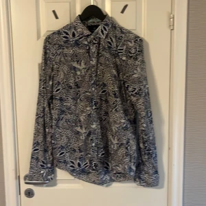 Mönstrad blå och vit skjorta - Säljer en snygg långärmad skjorta med blått och vitt blommigt mönster. Skjortan har klassisk krage och knäppning framtill. Perfekt för dig som vill sticka ut lite extra med en unik stil.