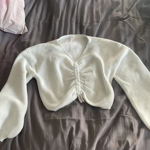 Vit ribbad croppad tröja med knyt - Säljer en vit ribbad croppad tröja med v-ringning och knyt framtill. Tröjan har långa ärmar och är gjord i ett mjukt stickat material. Perfekt för lager-på-lager och enkel att styla till olika outfits.