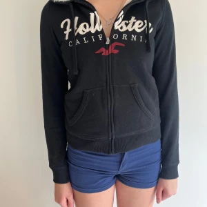 Svart hoodiejacka från Hollister - Mysig svart hoodiejacka från Hollister med vit fluffig foder på insidan. Jackan har dragkedja, huva med dragsko och två fickor framtill. Stor logga och text på bröstet i vitt och rött. Perfekt för kyliga dagar.