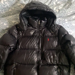 Svart dunjacka från Polo Ralph Lauren - Säljer en svart dunjacka från Polo Ralph Lauren med huva och dragkedja. Jackan har en klassisk pufferstil och är dekorerad med den ikoniska röda broderade loggan på bröstet. Perfekt för kalla dagar och riktigt snygg till streetwear. Bra skick och knappt använd köpte jackan för 6000kr