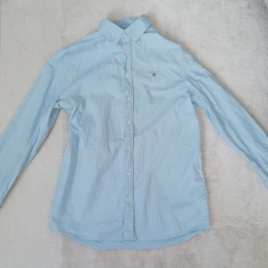 Ljusblå skjorta från GANT - Snygg ljusblå skjorta från GANT med klassisk button-down krage och broderad logga på bröstet. Skjortan har långa ärmar och knäppning framtill. Perfekt för en stilren look.