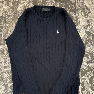 Mörkblå kabelstickad tröja från Polo Ralph Lauren - Säljer en klassisk mörkblå kabelstickad tröja från Polo Ralph Lauren med rund halsringning och broderad logga på bröstet. Tröjan har långa ärmar och ribbade muddar vid ärmslut och nederkant. Perfekt för lager-på-lager-stil.