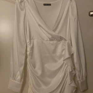 Vit omlottklänning med volang - Säljer en vit omlottklänning med långa ärmar och volangdetaljer längs sidan och nederkanten. Klänningen har v-ringning och är tillverkad i ett glansigt material som ger en elegant känsla.