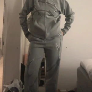 Grå mjukisbyxor med silvriga detaljer - Gråa mjukisbyxor med silvriga, blanka mönster längs benen. Byxorna har en avslappnad passform och resår i midjan. Perfekta för en chill och sportig stil. Byxorna är i storlek M och tröjan är i storlek S