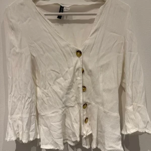 Vit blus med volang och knappar från H&M - Säljer en vit blus från H&M med v-ringning och dekorativa bruna knappar framtill. Blusen har långa ärmar med volang vid ärmslut och nederkant. Perfekt för en stilren och feminin look.