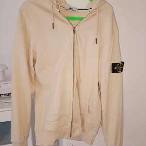 Beige hoodie från Stone Island - Säljer en beige hoodie från Stone Island med dragkedja och huva. Klassisk logga på vänster ärm och två fickor framtill. Perfekt för en avslappnad och stilren look.