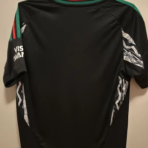 Arsenal svart bortatröja från Adidas - Snygg svart Arsenal bortatröja från Adidas med gröna och röda detaljer på axlarna. Tröjan har korta ärmar, klubbmärke och Adidas-logga på bröstet samt sponsortryck 'Emirates Fly Better' framtill.