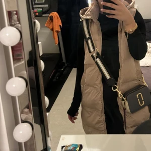 Beige lång quiltad väst med huva - Säljer en beige lång quiltad väst med huva och fickor. Perfekt att slänga över en hoodie eller tröja för extra värme. Västen har en avslappnad passform och stängs med dragkedja framtill.