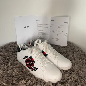 Gucci Ace Sneaker - Säljer ett par vita Gucci Ace sneakers med den ikoniska ormbroderingen på sidan och gröna samt röda detaljer. Skorna har snörning, rund tå och platt sula. Baktill finns metallicröda och gröna hälpaneler med Gucci-logga. Perfekta för dig som vill sticka ut. Tyvärr inget og kvar men kvitto finns. Nästintill nytt skick