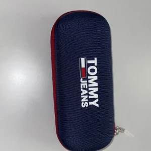 Mörkblå solglasögon från Tommy Jeans - Snygga mörkblå solglasögon från Tommy Jeans med klassisk rektangulär form. Kommer i ett matchande fodral med röd och blå design. Perfekta för soliga dagar och har diskret logga på skalmen.