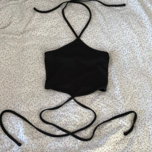 Svart ribbad croppad linnetopp från Amisu - Säljer en svart ribbad croppad linnetopp från Amisu med smala knytband som knyts i nacken och runt midjan. Perfekt för sommaren och enkel att styla till olika outfits. Materialet är mjukt och stretchigt.