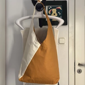 Tvåfärgad tygväska i beige och orange - Säljer en stor och rymlig tygväska i beige och orange. Väskan har en enkel och stilren design med bred axelrem och är perfekt för att bära med sig allt man behöver under dagen. Passar lika bra till skolan som till stan.