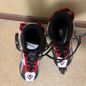 Svarta och röda inlines stl 33-36 - Säljer ett par snygga inlines från Pros i svart, rött och vitt. Skorna har snörning och spänne för extra bra passform. Perfekta för dig som gillar att åka snabbt och smidigt på asfalt.