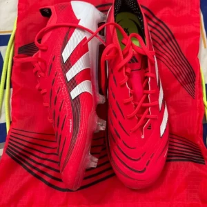 Röda Adidas Predator fotbollsskor - Säljer ett par röda Elite Adidas Predator fotbollsskor med vita och svarta detaljer. Skorna har snörning, mönstrad ovandel och dobbar för spel på gräs. Ikonisk Predator-logga under sulan och klassiska tre ränder på sidan.