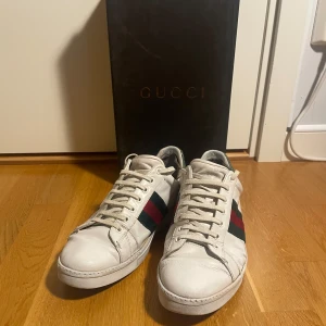 Gucci ace skor - Snygga vita sneakers från Gucci med klassiska ränder i grönt och rött på sidan. Skorna har snörning och är tillverkade i skinn, vilket ger en lyxig känsla. Perfekta för dig som vill ha en stilren och exklusiv look. Skornas skick är cirka 6/10. OG box medföljer. Hör gärna av dig om du undrar något! Storlek 40,5 men passar som 42