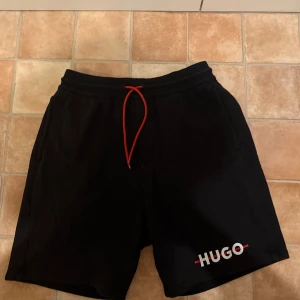 Svarta shorts från HUGO med röd snörning - Svarta shorts från HUGO med vit logga på vänster ben och röd snörning i midjan. Klassisk passform med elastisk midja och fickor på sidorna samt en bakficka. Perfekta för en avslappnad stil.