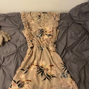 Beige blommig klänning med spets och volang - Söt beige klänning med blommigt mönster, volangdetaljer längs axlarna och spets upptill både fram och bak. V-ringad och med knappar i ryggen. Perfekt för somriga dagar.