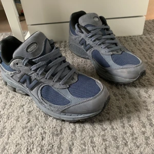 Grå blå New Balance 2002 r - Säljer ett par grå New Balance sneakers med blå meshpaneler. Skorna har rund tå, platt sula och klassisk snörning. Mycket bra skick, knappt använda. Storlek 40,5