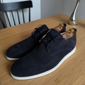 Mörkblå mocka loafers med vit sula - Snygga mörkblå loafers i mocka med vit platt sula och klassisk snörning. Skorna har en stilren design och rund tå, perfekta för en clean och modern look. Använd endast ett fåtal gånger.