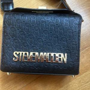 Svart axelväska från Steve Madden - Säljer en svart axelväska från Steve Madden med präglat S-mönster och stor guldfärgad logotyp framtill. Väskan har flera fack inuti och stängs med ett snyggt guldfärgat spänne. Perfekt för att bära dina viktigaste saker med stil.