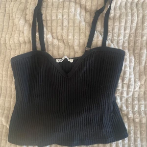Svart ribbad linnetopp från Zara - Säljer en svart ribbad linnetopp från Zara med justerbara axelband och v-ringad skärning. Perfekt att styla till olika outfits och enkel att matcha med både jeans och kjol.