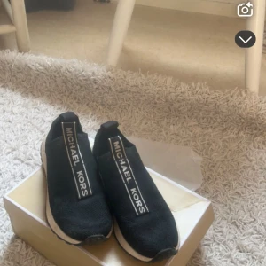 Svarta slip-on sneakers från Michael Kors - Snygga svarta slip-on sneakers från Michael Kors med tydlig logga på ovansidan. Skorna har en sportig design med vit sula och är perfekta för en trendig vardagslook.