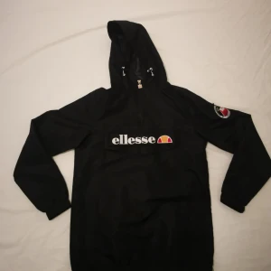 Svart vindjacka från Ellesse - Säljer en svart vindjacka från Ellesse med huva och dragkedja. Jackan har stor logga på bröstet och en patch på ärmen. Perfekt för blåsiga dagar och enkel att matcha med olika outfits.