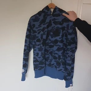 Blå camo hoodie från A Bathing Ape(bape) - Säljer en blå hoodie från Bape med svart camouflagemönster och ikonisk hajdesign på huvan. Tröjan har dragkedja och ribbade muddar. Perfekt för dig som gillar streetwear. Storlek XL men liten I storlek så funkar bäst för L
