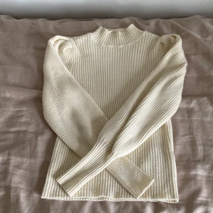 Ribbad krämvit stickad tröja från ONLY - Säljer en ribbad, krämvit stickad tröja från ONLY med puffade axlar och hög hals. Tröjan har långa ärmar och en figurnära passform. Perfekt för lager-på-lager och stilrena outfits.
