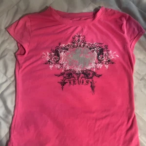 Rosa t-shirt med tryck y2k - Säljer en rosa t-shirt med korta ärmar och ett stort dekorativt tryck i svart, vitt och grått framtill. T-shirten har en klassisk passform och är tillverkad i mjuk bomull. Den passar till dig som gillar y2k stilen