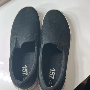 Svarta slip-on sneakers med vit sula - Svarta slip-on sneakers med vit, platt gummisula och rund tå. Skorna är i textilmaterial och har en enkel, stilren design utan snörning eller extra detaljer. Perfekta för en avslappnad och bekväm stil.