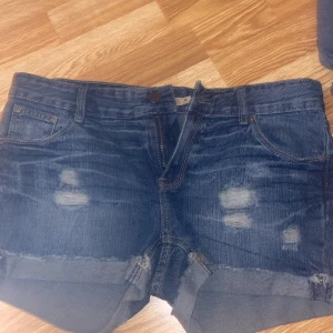 Blå jeansshorts från Serious Sally - Snygga blå jeansshorts från Serious Sally med slitna detaljer och uppvikta benslut. Klassisk femficksmodell och knappgylf. Perfekta för en avslappnad stil.
